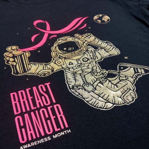 A shirt that says "Breast Cancer Awareness Month" with an astronaut with a spray can spraying a pink ribbon